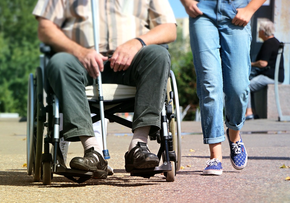 En person sitter i rullstol och håller i en käpp med en stödperson i blå jeans som går bredvid