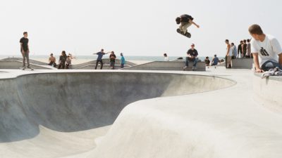 En skateboardpark vid havet. En person åker i en bowl och hoppar högt upp i luften med brädan. Runt om står människor och tittar på.